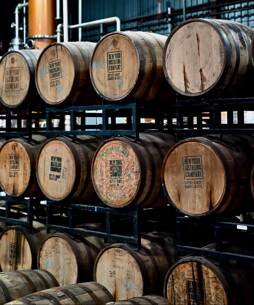 Multiple wooden whiskey barrels stacked on black metal racks inside a distillery or warehouse, with visible staves, metal hoops, and printed barrel labels.