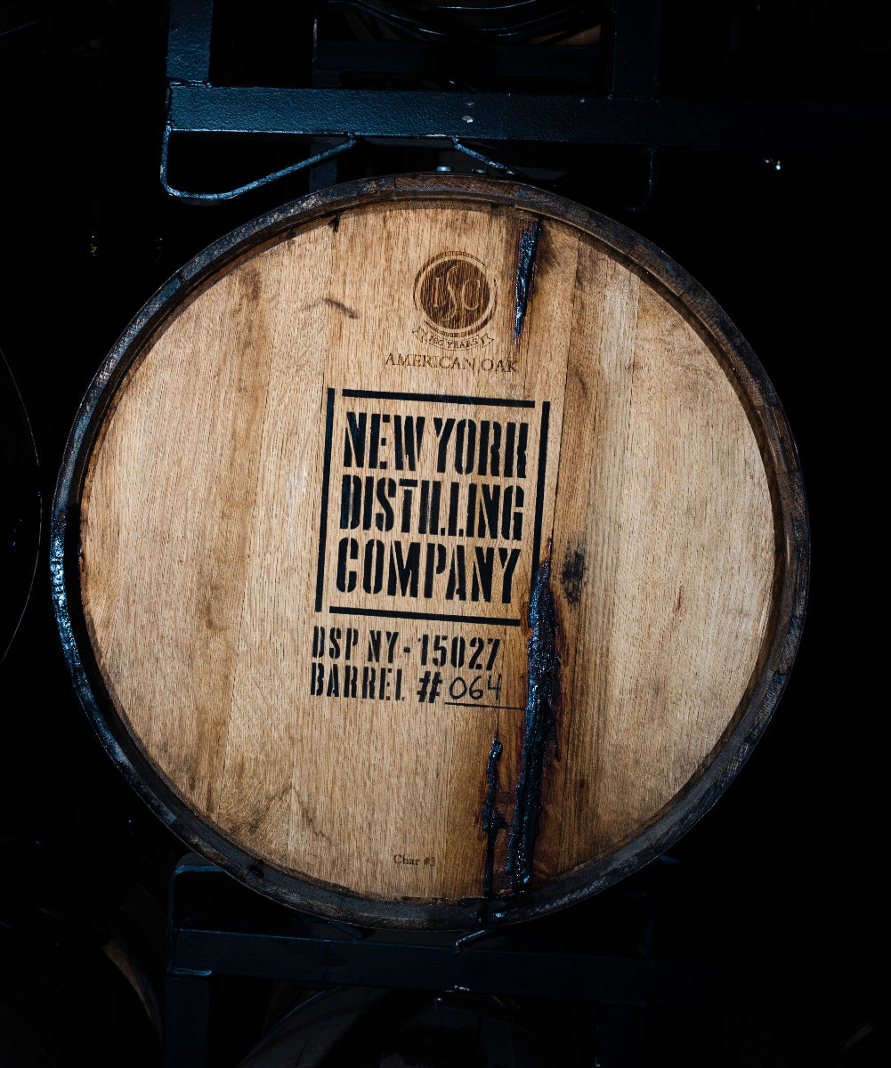 A close-up of a wooden whiskey barrel stave with black printed text reading 'New York Distilling Company' and 'BSP NY-15027 Barrel #25', photographed in a dark distillery setting with other barrels partially visible.